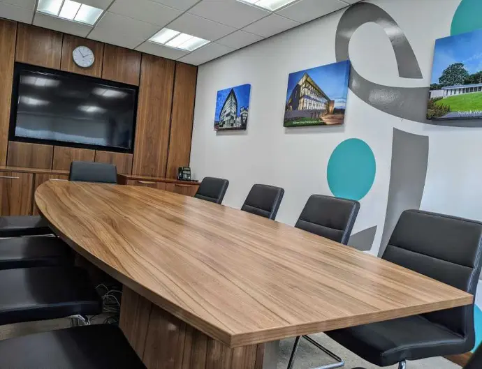 A conference room available for hire in Dundee featuring a long wooden table, black chairs, a large wall-mounted TV, a clock, and framed pictures on the wall.