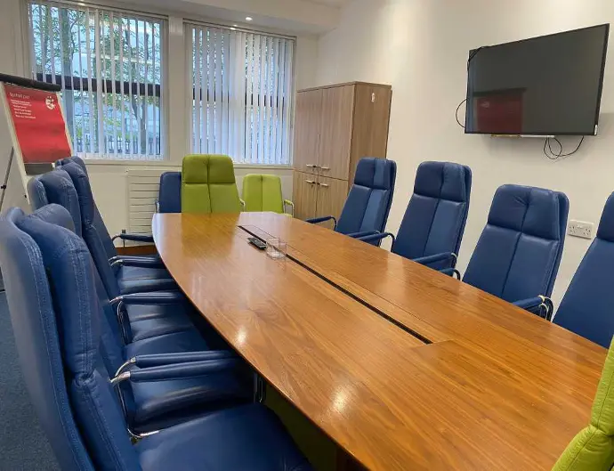 A meeting room available for hire in Dundee featuring a long wooden table, blue and green chairs, a wall-mounted TV, and a large window.