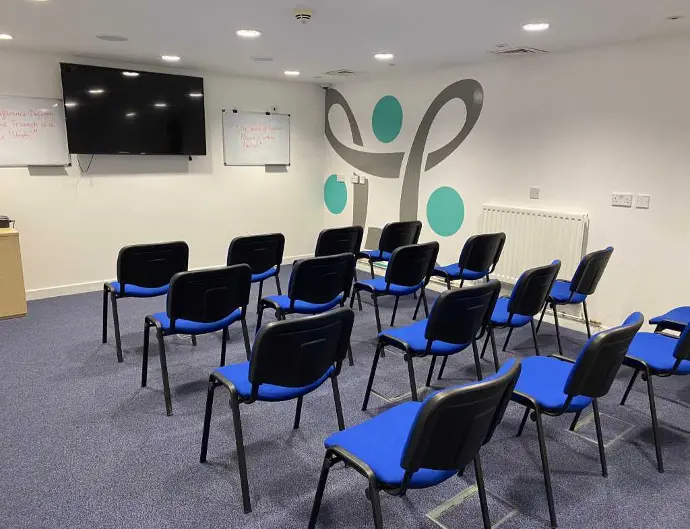 A training room to hire in Dundee featuring rows of blue chairs facing a mounted TV screen, whiteboards, and a wall with decorative designs.