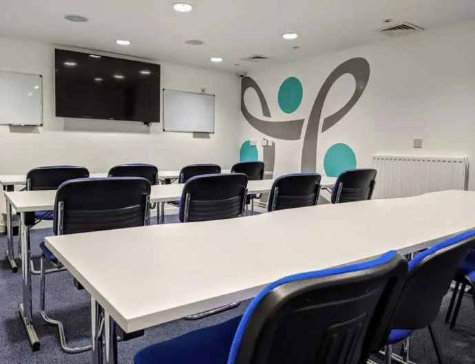 A training room to hire in Dundee featuring rows of blue chairs facing a mounted TV screen, whiteboards, and a wall with decorative designs.